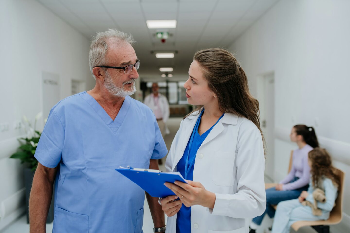 Older doctor giving advise to his younger colleague, discussing at hospital corridor. Health care